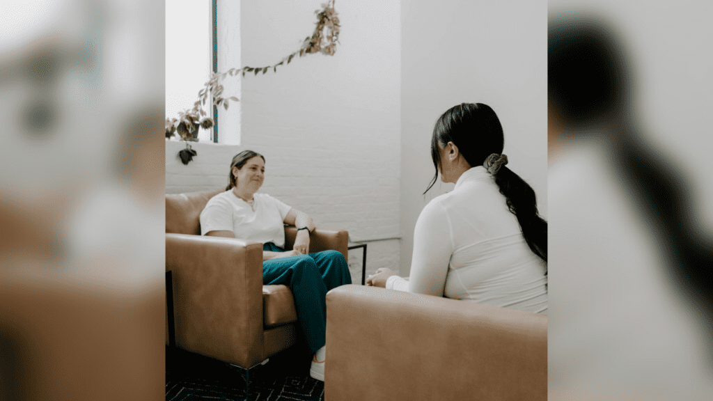 A female therapist in a white shirt sitting in a brown chair smiling at a female patient.