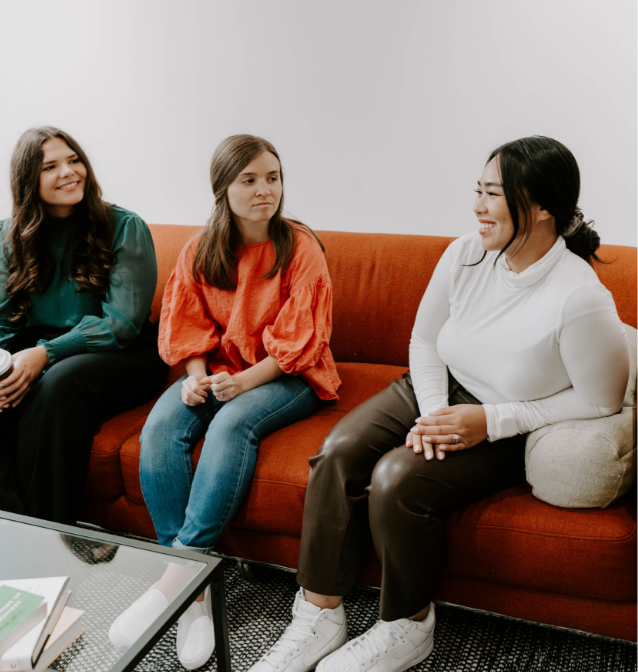 Three female counselors sitting in a therapy office on an orange couch having a discussion.