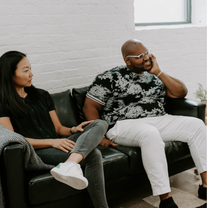 A black male mental health counselor sitting on a black leather couch in an office next to an Asian female counselor.