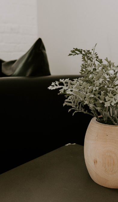 A decorative plant in a wooden vase on a side table next to a black leather couch inside a therapist office.