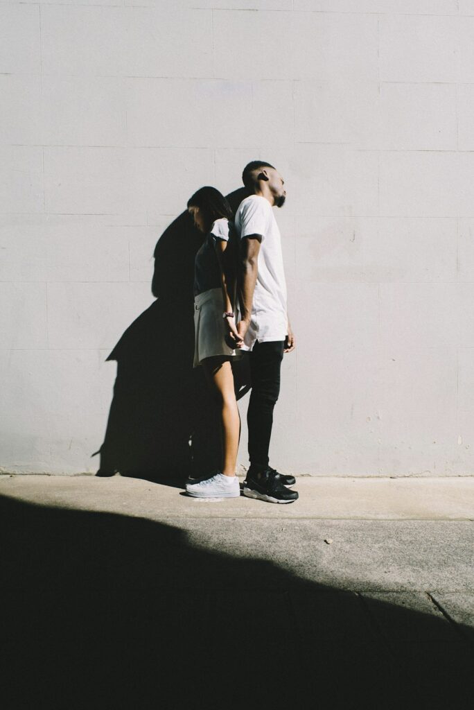 multiracial couple standing back to back holding hands next to white wall