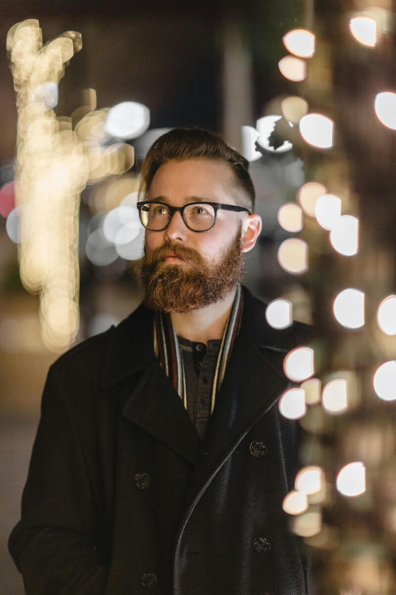 Man with red beard and glasses standing behind tree with Christmas lights
