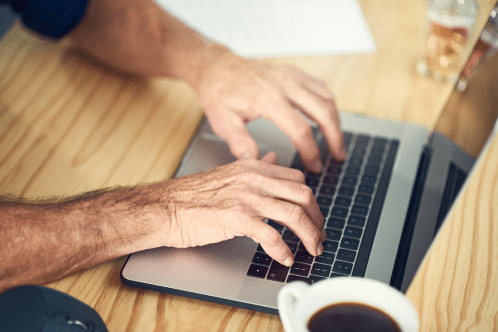 A young man typing on a silver laptop to do research on his Self-Diagnosis, with a cup of coffee on a tan wooden background.