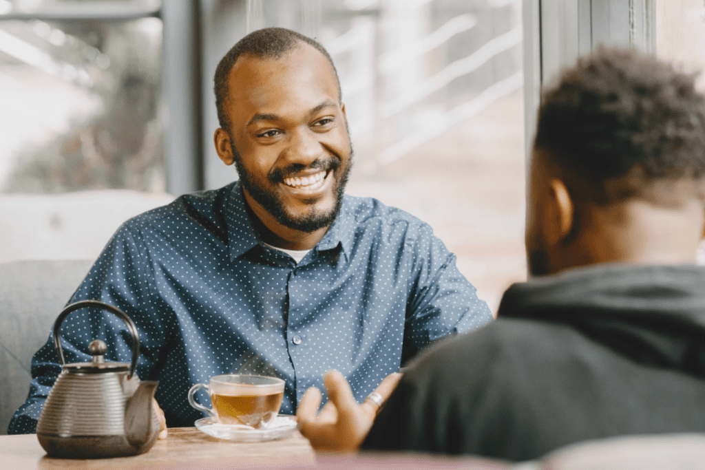 A young man with a dark blue button down and a young man in a black hoodie discussing Self-Diagnosis over a cup of tea.