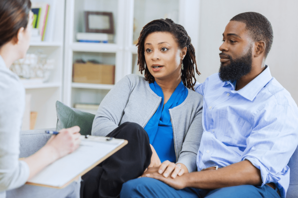 Young woman with brown hair wearing grey sweater and blue blouse with young man with black hair, wearing a blue button down, in couples therapy, with their young woman therapist with brown hair wearing a white sweater.
