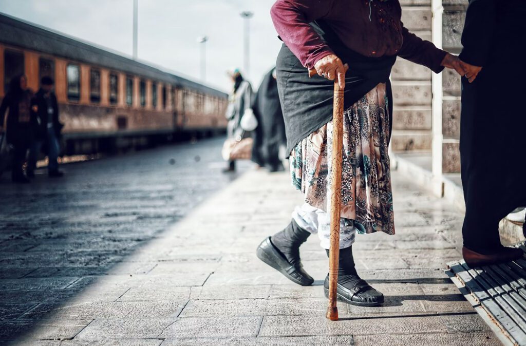Elderly lady with a fuchsia sweater and cane being assisted by a kind Samaritan at a train station.