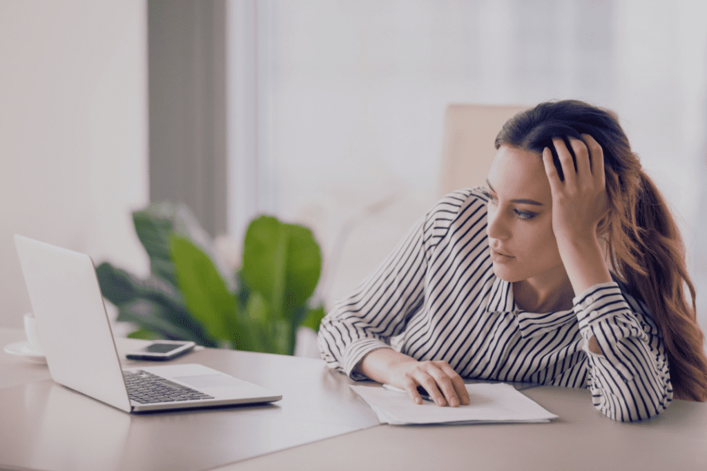 Young woman with long brown hair wearing a white and black striped button down, sitting at her white work desk, staring at her computer in front of her bored and tiredly, with her hand on her head, leaning against the desk, with a big green plant and white curtains in the background.