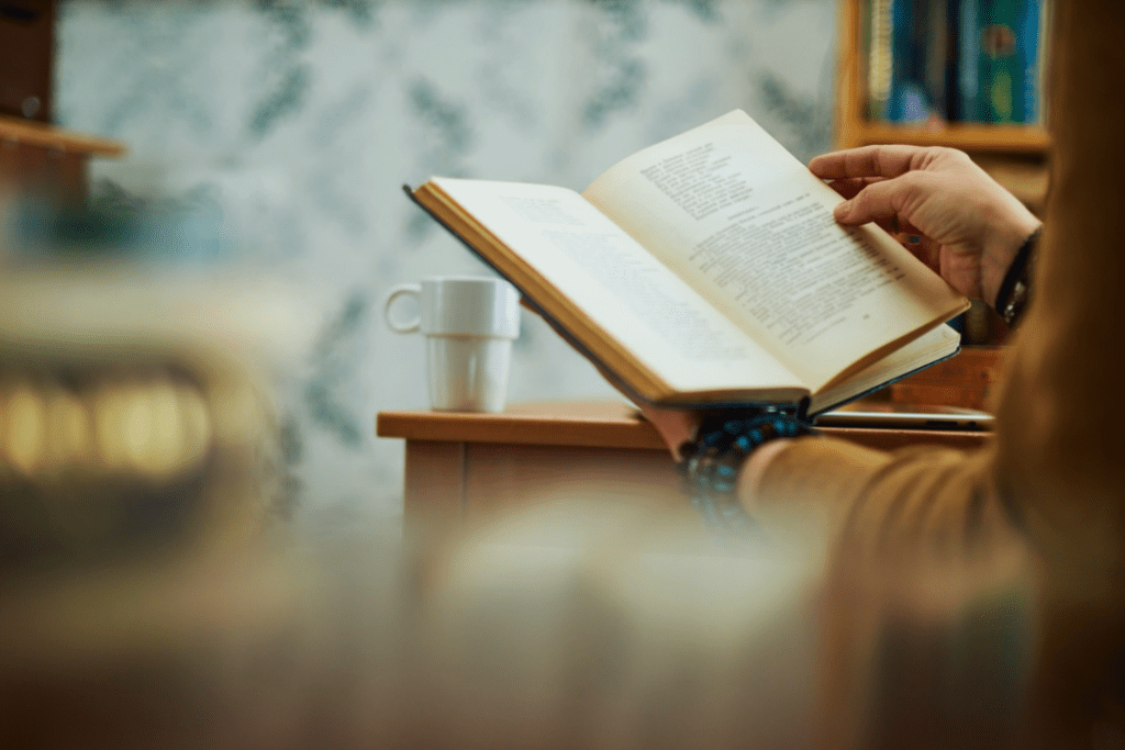 Young woman wearing a tan sweater and dark blue and black bracelets on her left hand, reading a book, with a cup of coffee on her tan wooden desk, taking time away from electronics. 