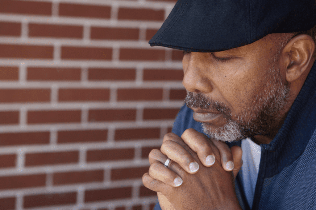 Older man wearing a black hat and a blue and white t-shirt, with his hands held together, reflecting on his loss.