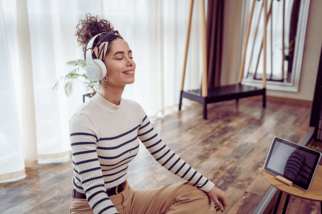 A young woman with her eyes closed, with brown hair, wearing a white and black striped shirt and khaki pants, wearing white wireless over the ear headphones, sitting on a brown wooden floor, following a meditation exercise on her tablet on her end table, practicing mindfulness, with a white background of white curtains in her home. 