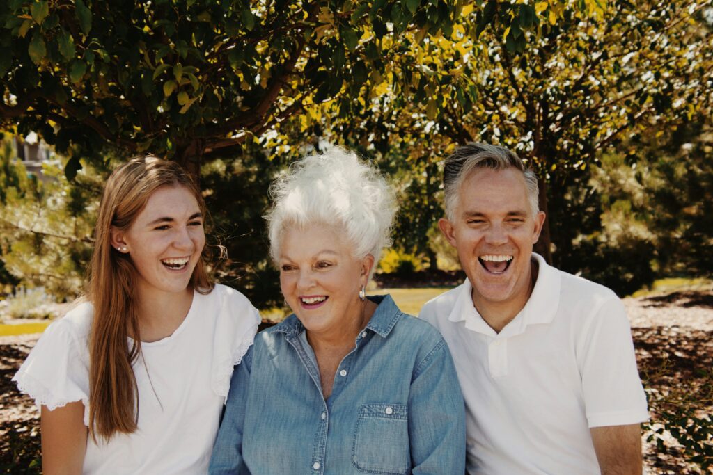 Three people of different generations laughing together outdoors under a tree, showing the joy and connection that can exist across age groups
