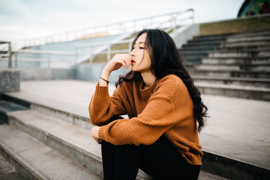 Woman wearing orange sweater and black pants sitting outside thinking.
