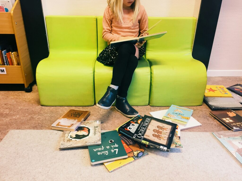 Preteen girl sitting on a green couch reading a book with a pile of children’s books on the floor around her