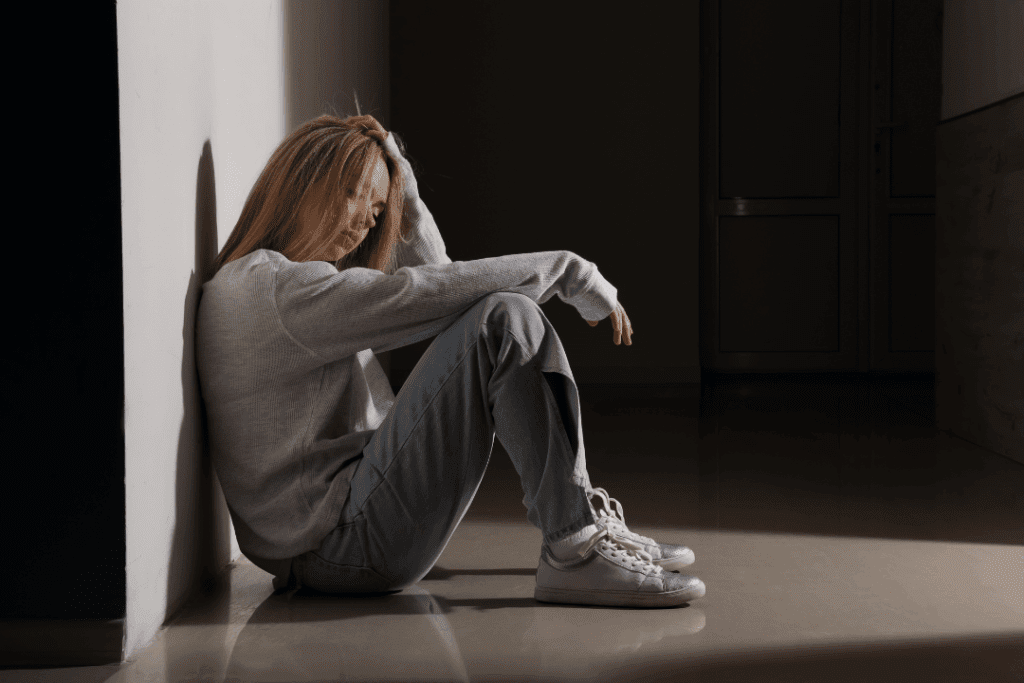A young woman sitting on the floor against a wall, her posture slumped and her head resting in her hand, suggesting she’s feeling overwhelmed or emotionally drained. The dim lighting and empty hallway around her emphasize a sense of loneliness or heaviness, as if she’s taking a moment to pause, breathe, or process something difficult.
